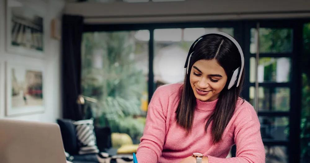 Woman wearing headphones studying and writing in a notebook beside a laptop.