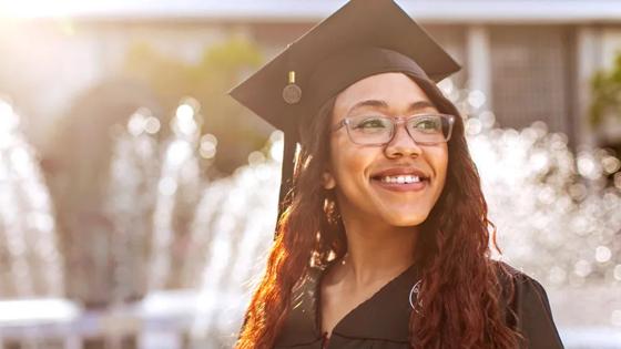 An ODU graduate wearing a graduation cap and gown smiles in the sunlight near ODU's lion fountain.
