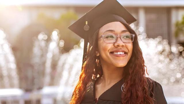 An ODU graduate wearing a graduation cap and gown smiles in the sunlight near ODU's lion fountain.