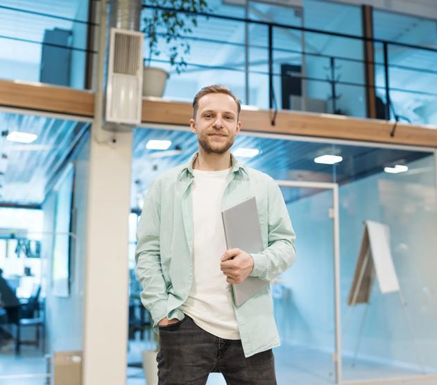 Confident male entrepreneur looking at camera standing in modern office hall. Young man in casual holding laptop computer, his hand in pocket