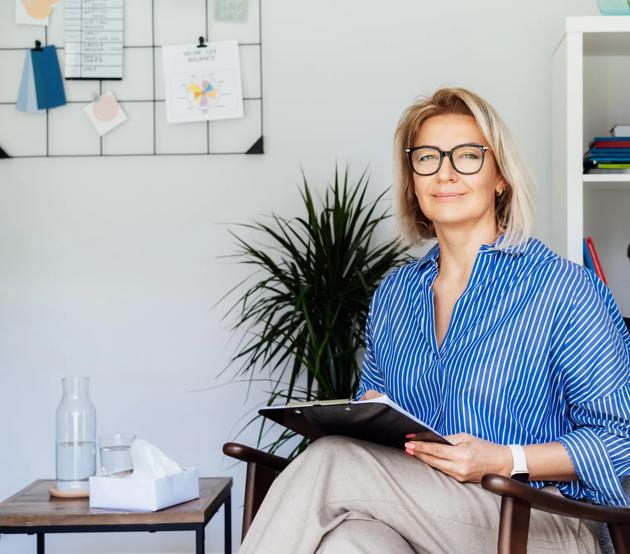 A middle aged woman sits holding clipboard and smiling at the camera