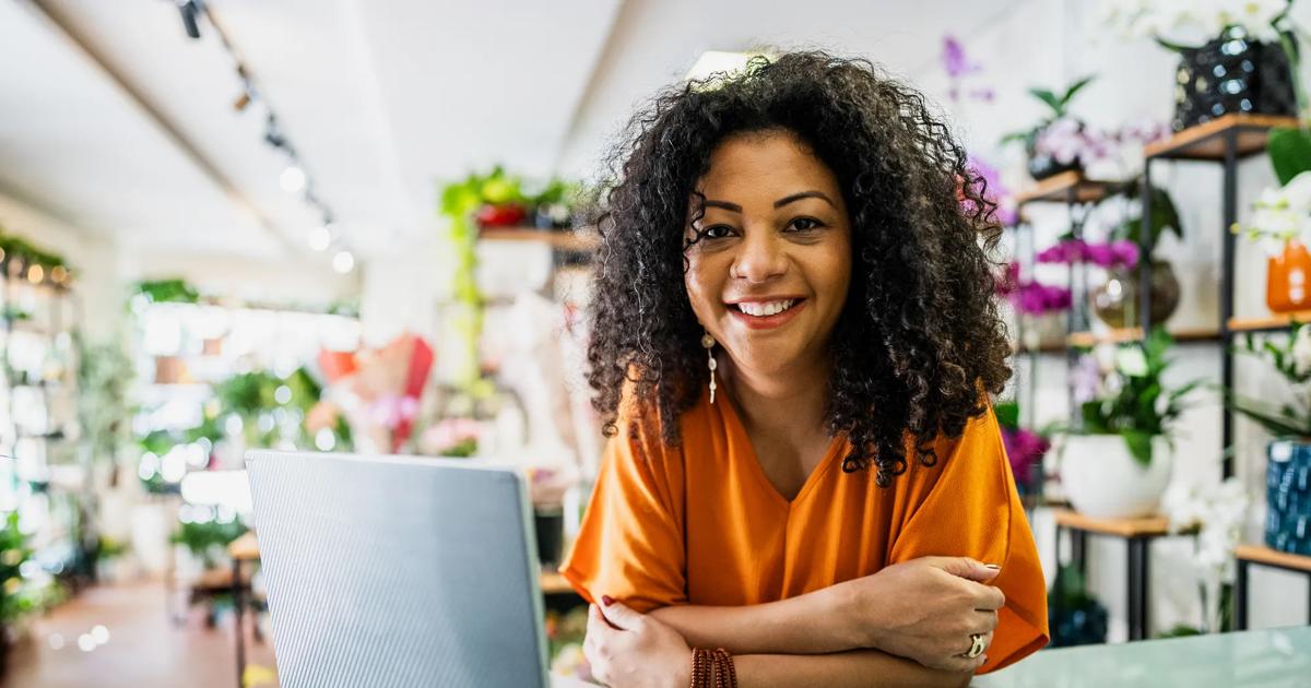 A woman wearing an orange shirt smiles at the camera leans on a desk in front of a laptop at her small flower business.