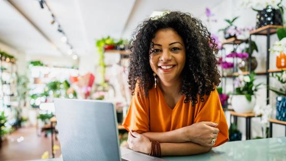 A woman wearing an orange shirt smiles at the camera leans on a desk in front of a laptop at her small flower business.
