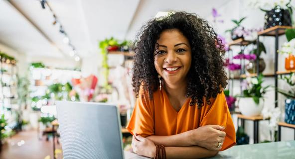 A woman wearing an orange shirt smiles at the camera leans on a desk in front of a laptop at her small flower business.