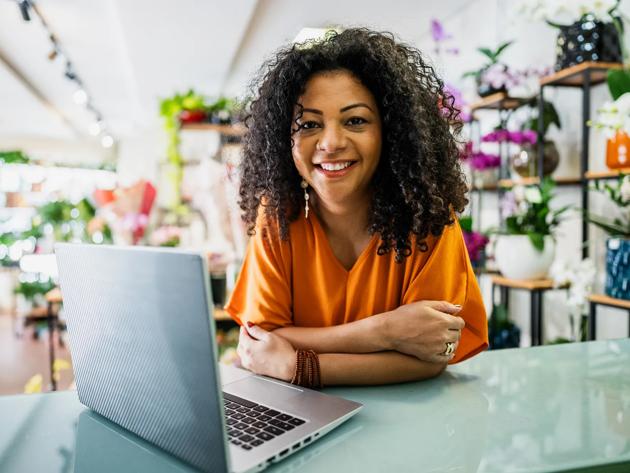 A woman wearing an orange shirt smiles at the camera leans on a desk in front of a laptop at her small flower business.