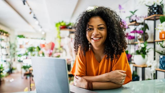 A woman wearing an orange shirt smiles at the camera leans on a desk in front of a laptop at her small flower business.