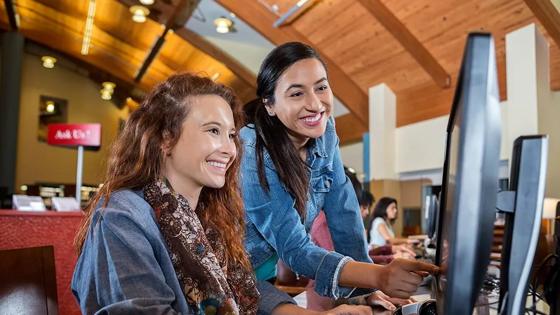 A female librarian guides a young student to find information at a desktop computer.