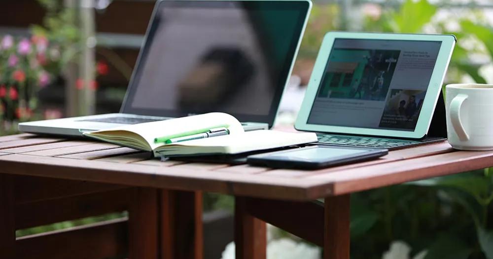 Laptop, tablet, notebook, and coffee on a desk in a bright home study space.