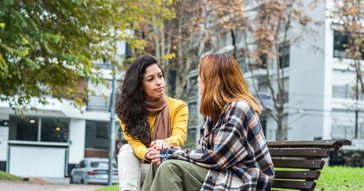 A woman sits outside on a public bench with another person facing away from the camera, holding hands.