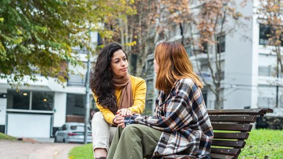 A woman sits outside on a public bench with another person facing away from the camera, holding hands.