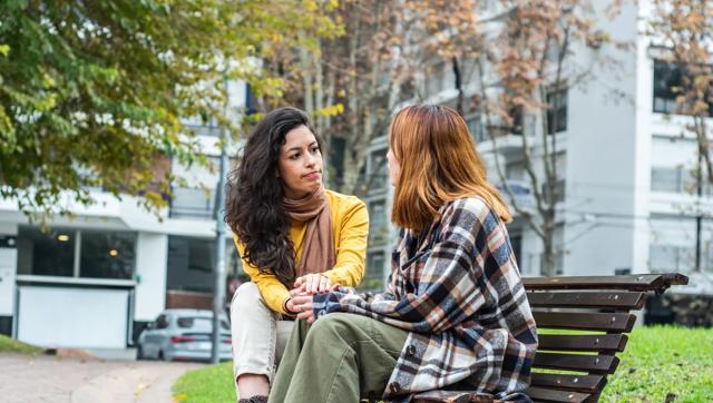 A woman sits outside on a public bench with another person facing away from the camera, holding hands.
