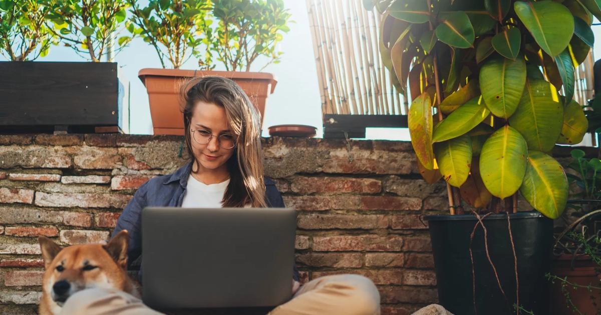 Positive young millennial girl with long hair sitting outdoor at home terrace with best friend dog working on distance using laptop, beautiful student girl having online video meeting via computer